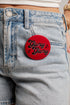 A close-up of a person wearing light-wash denim shorts and a white cropped top, with a round red pin on the back of the shorts that reads &