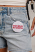 A close-up of a person wearing light-wash denim shorts and a white top, with a round white pin or badge attached to the pocket. The badge has red text that reads “SATURDAY IN ATHENS.