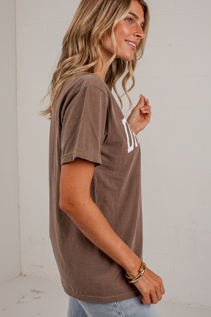 A woman wearing a brown short-sleeve t-shirt with a white graphic print on the chest, paired with light blue jeans, standing against a white brick wall.