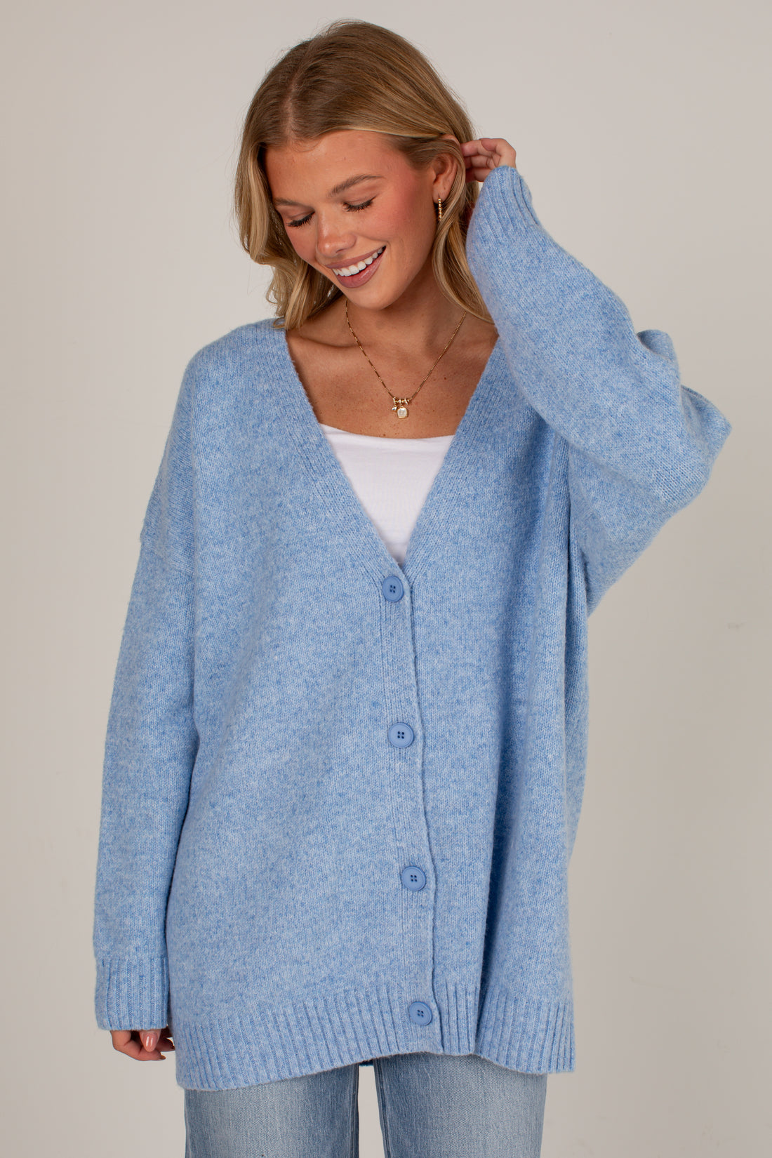A woman wearing an oversized light blue knit cardigan with a white V-neck undershirt and light blue jeans, photographed against a plain light beige studio background.