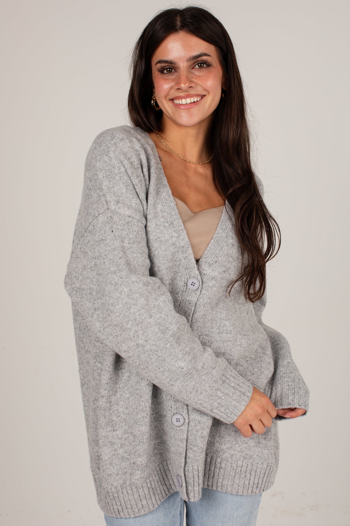 A woman wearing a light gray heather knit cardigan over a beige top and light blue jeans, standing against a plain light beige studio background.