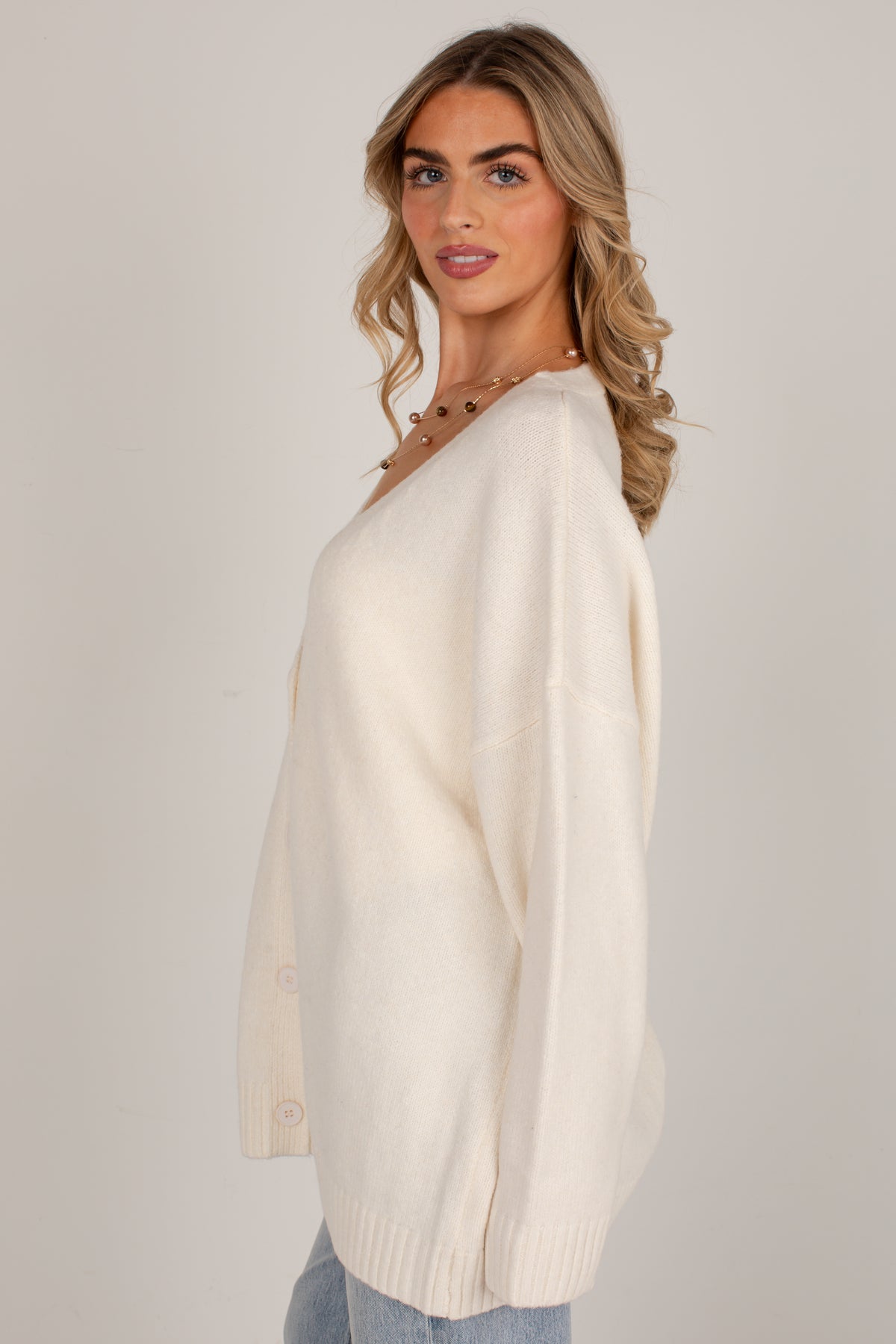 A woman wearing an oversized cream-colored knit sweater with a V-neck and long sleeves, paired with light blue jeans, photographed against a plain light gray studio background.