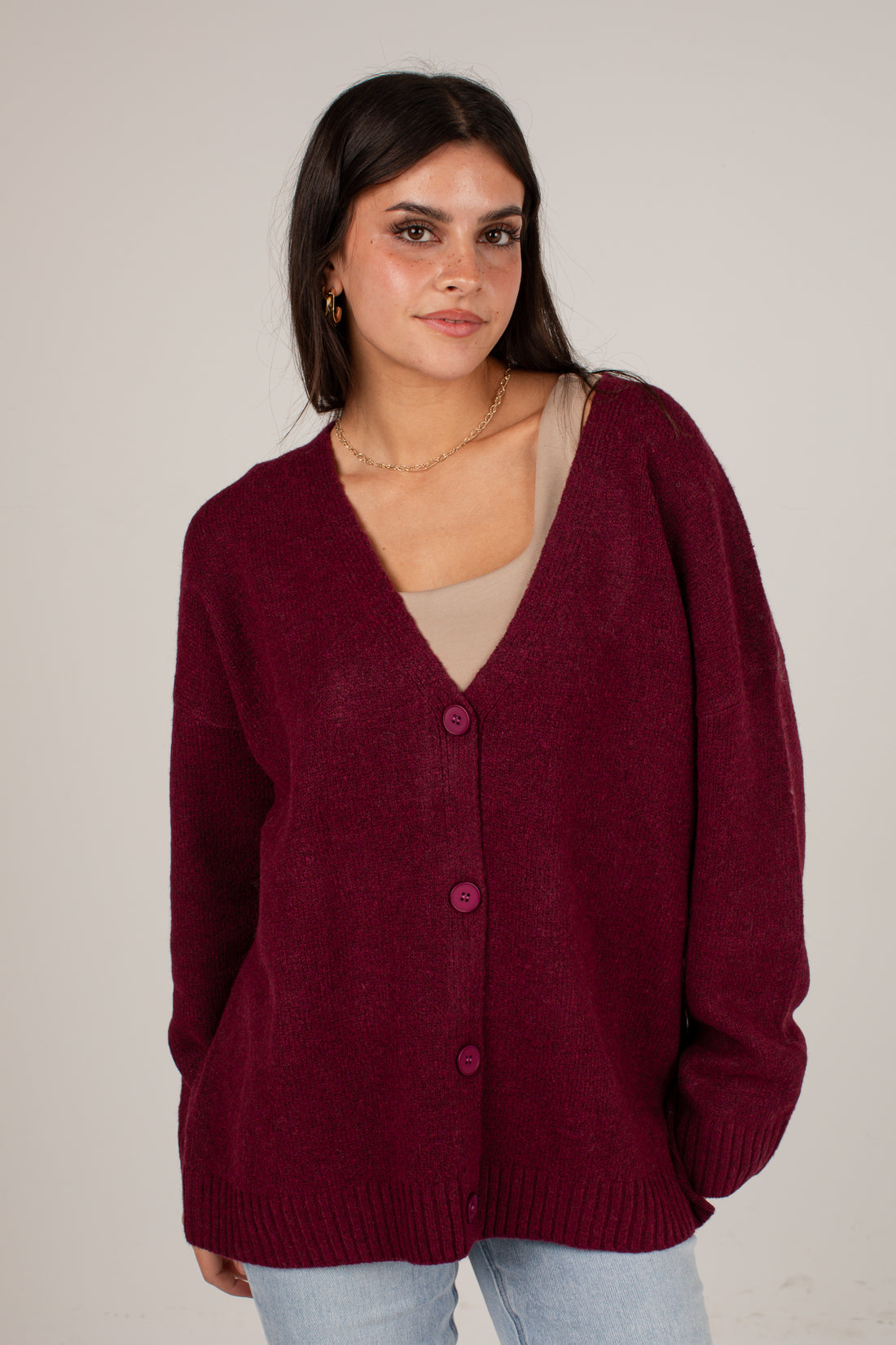 A woman wearing an oversized burgundy knit cardigan with a V-neck and front buttons, layered over a beige top and light blue jeans, photographed against a plain light gray studio background.