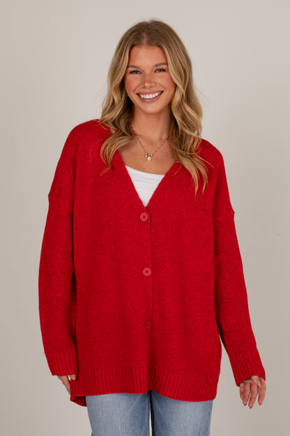 A smiling blonde woman wearing a bright red knit cardigan with three front buttons over a white top and light blue jeans, photographed against a plain light beige studio background.