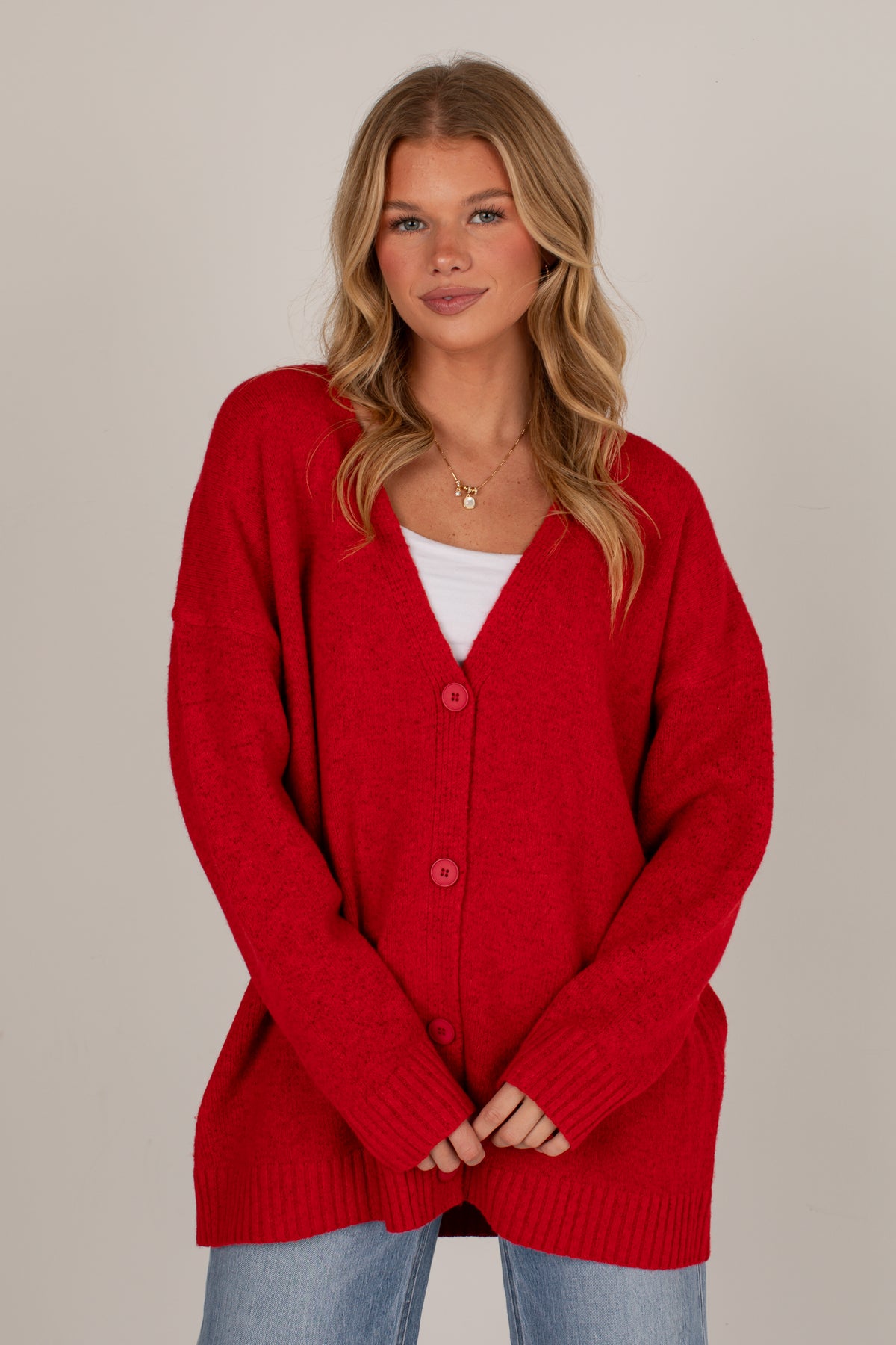 A woman wearing a bright red knit cardigan over a white top and light blue jeans, standing against a plain light gray studio background.