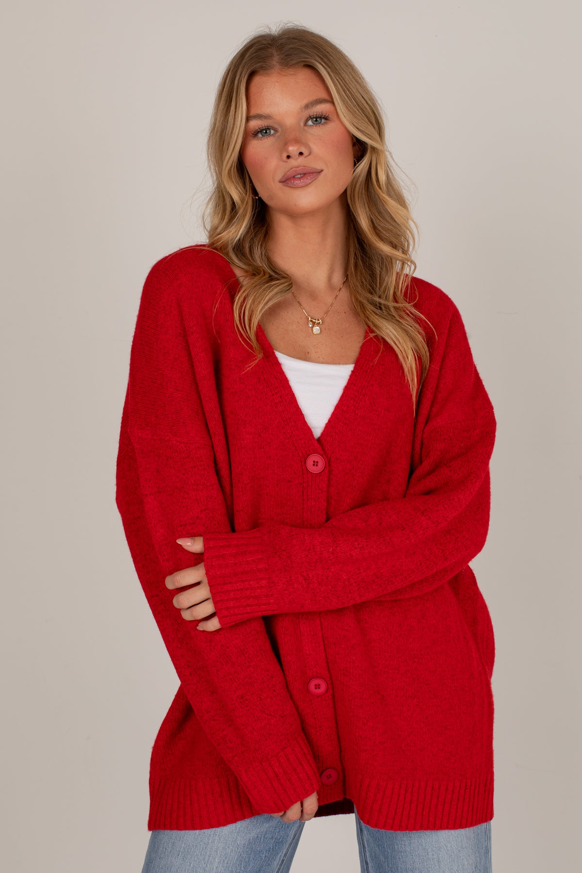 A woman wearing a bright red knit cardigan over a white top and light blue jeans, standing against a plain light gray studio background.
