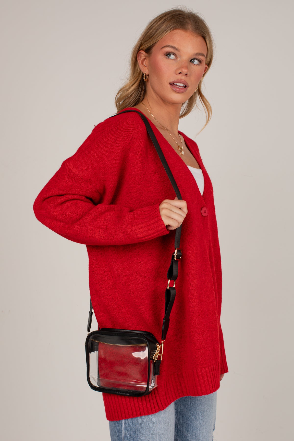 A woman wearing a bright red knit cardigan and light blue jeans, carrying a small black crossbody bag with a clear front pocket. She is photographed against a solid light gray studio background.