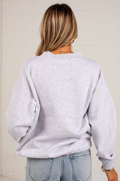 A woman viewed from behind wearing a light heather gray crewneck sweatshirt and light blue jeans, standing against a light-colored wooden wall.