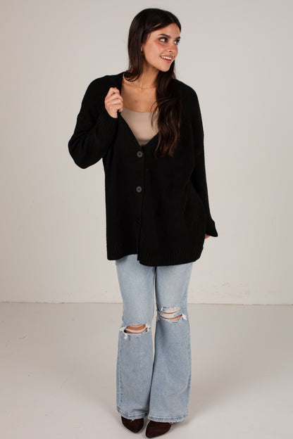 A woman modeling a black button-up knit cardigan over a beige top and light-wash distressed flare jeans, standing against a plain light gray studio background.