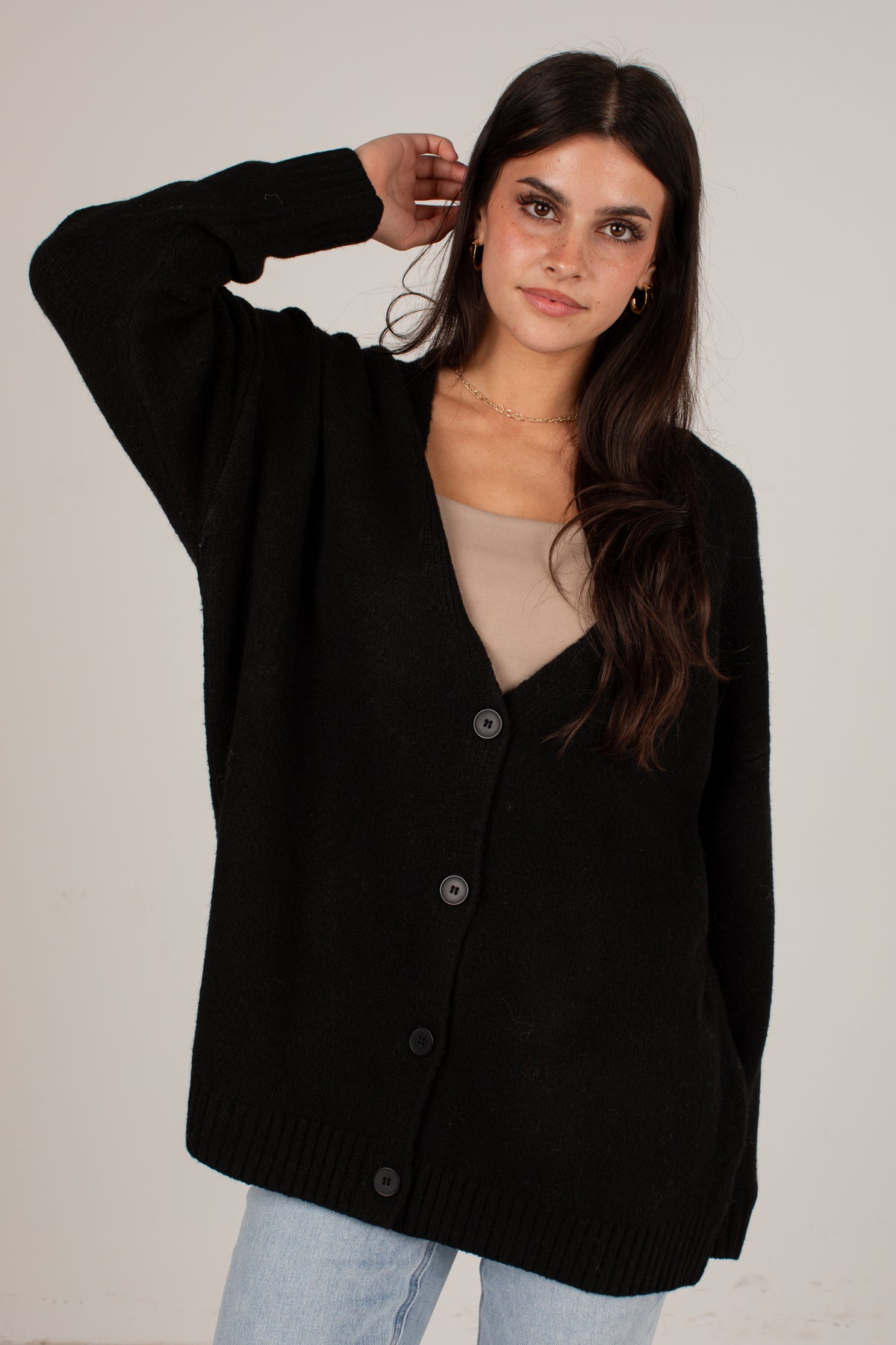 A woman wearing a black button-front cardigan over a beige top and light blue jeans, standing against a plain light background.