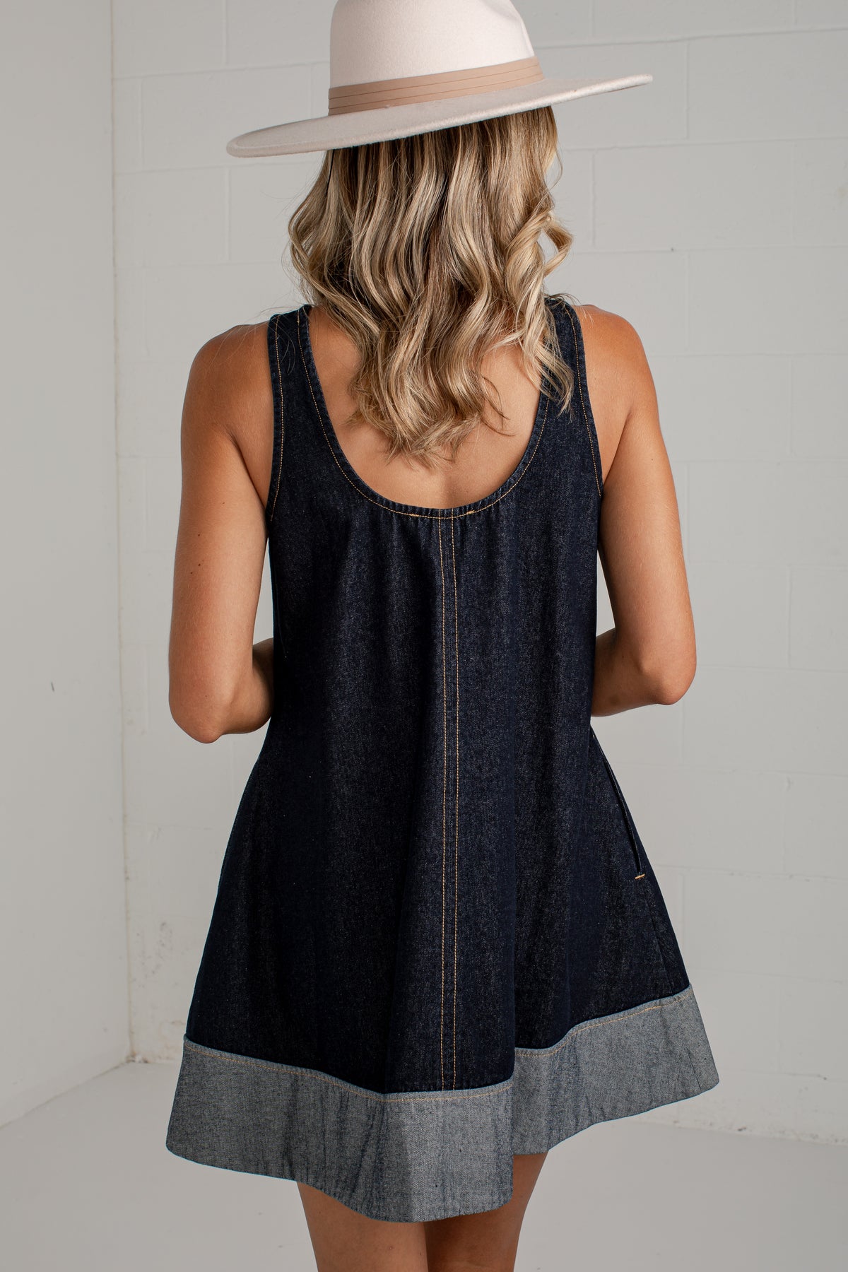 A woman viewed from behind wearing a sleeveless dark denim dress with a lighter gray cuffed hem and a wide-brim beige hat, standing against a white wall.