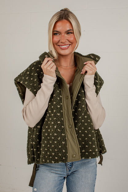 A smiling blonde woman wearing a sleeveless olive green vest with a beige and green pattern over a cream long-sleeve shirt and blue jeans, standing against a white brick wall.