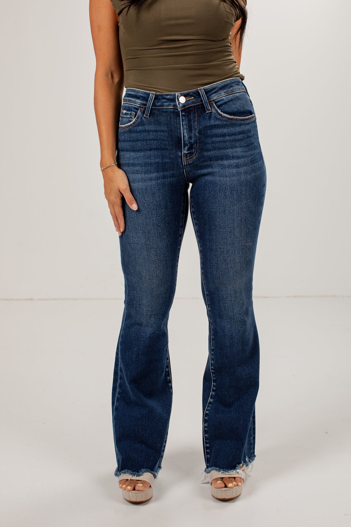 A model wearing dark blue high-waisted flare jeans with a frayed hem, paired with an olive green top and beige platform sandals, standing against a plain white studio background.