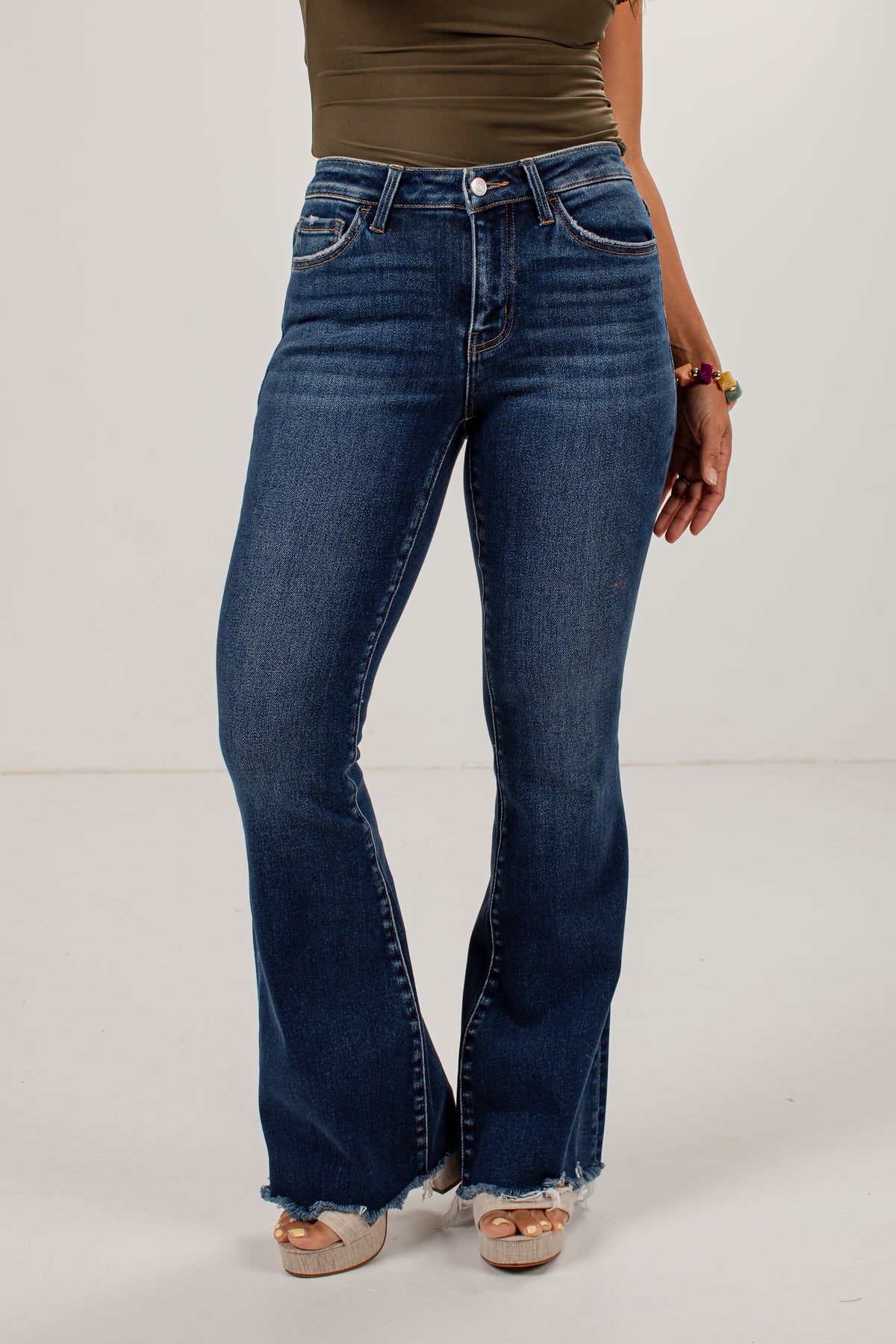 A model wearing dark blue high-waisted flare jeans with a frayed hem, paired with a brown top and beige platform sandals, shown against a plain white studio background.