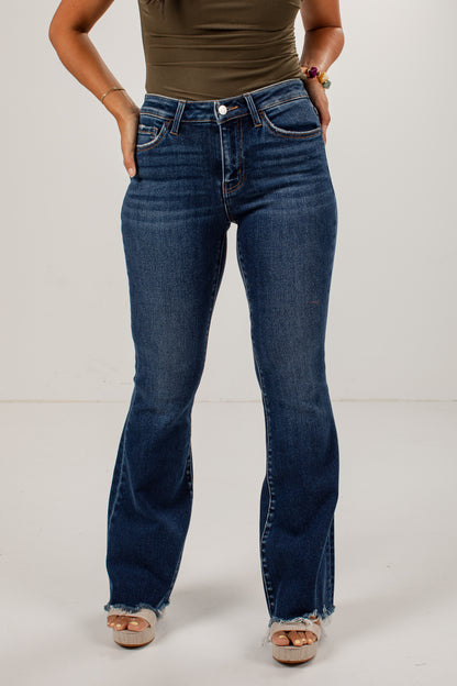A person modeling dark blue flare jeans with a frayed hem, paired with a brown top and beige platform sandals, standing against a plain light gray studio background.