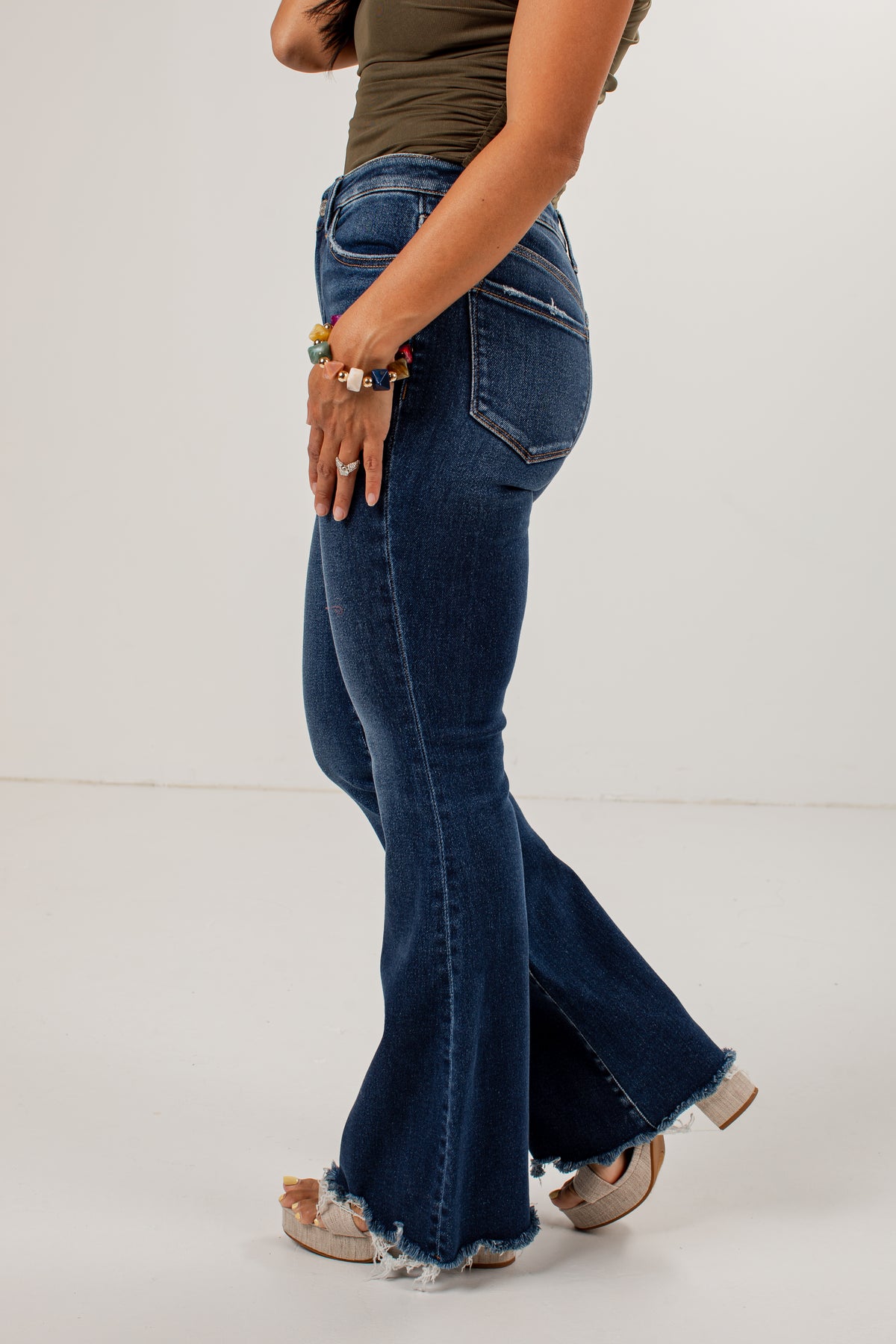 A side-profile view of a model wearing dark blue high-waisted flared jeans with a raw frayed hem, paired with a brown top and beige flat shoes, against a plain white studio background.