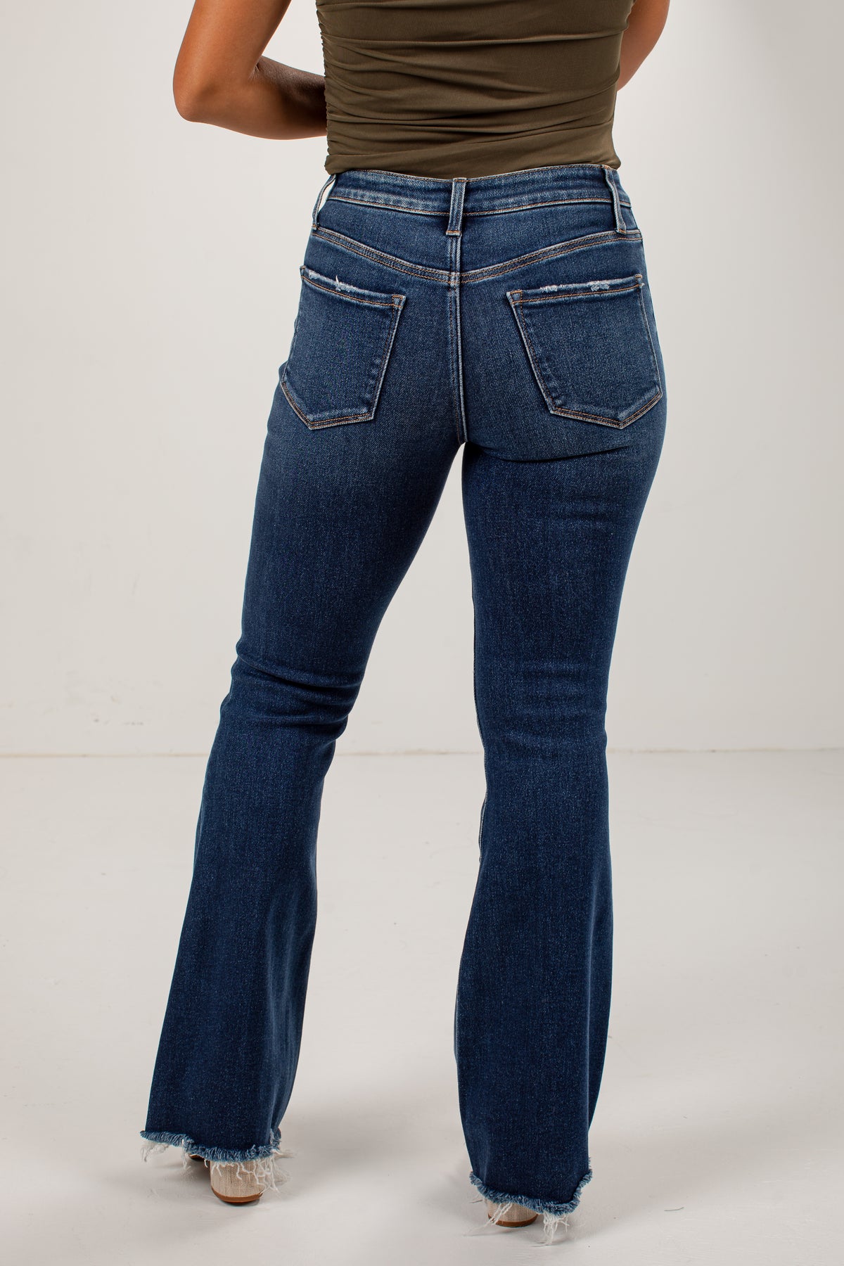 Rear view of a person wearing dark blue flare jeans with frayed hems and back pocket details, paired with an olive green top, against a plain light gray studio background.