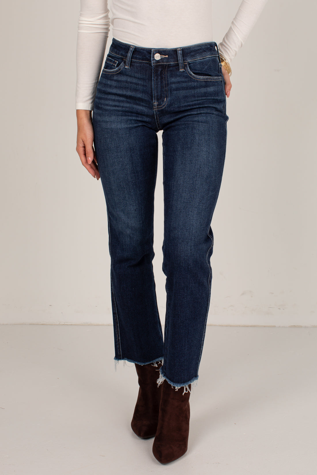 A model wearing dark blue straight-leg denim jeans with a raw, frayed hem, styled with a white long-sleeve top and brown suede ankle boots against a plain off-white studio background.