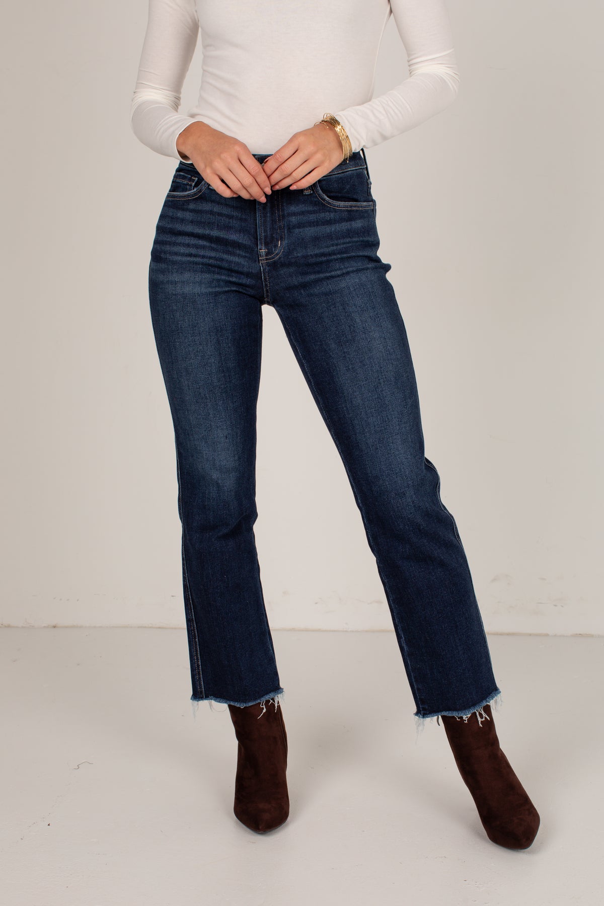 A model wearing dark blue straight-leg denim jeans with a raw frayed hem, styled with a white long-sleeve top and dark brown suede ankle boots against a plain off-white studio background.