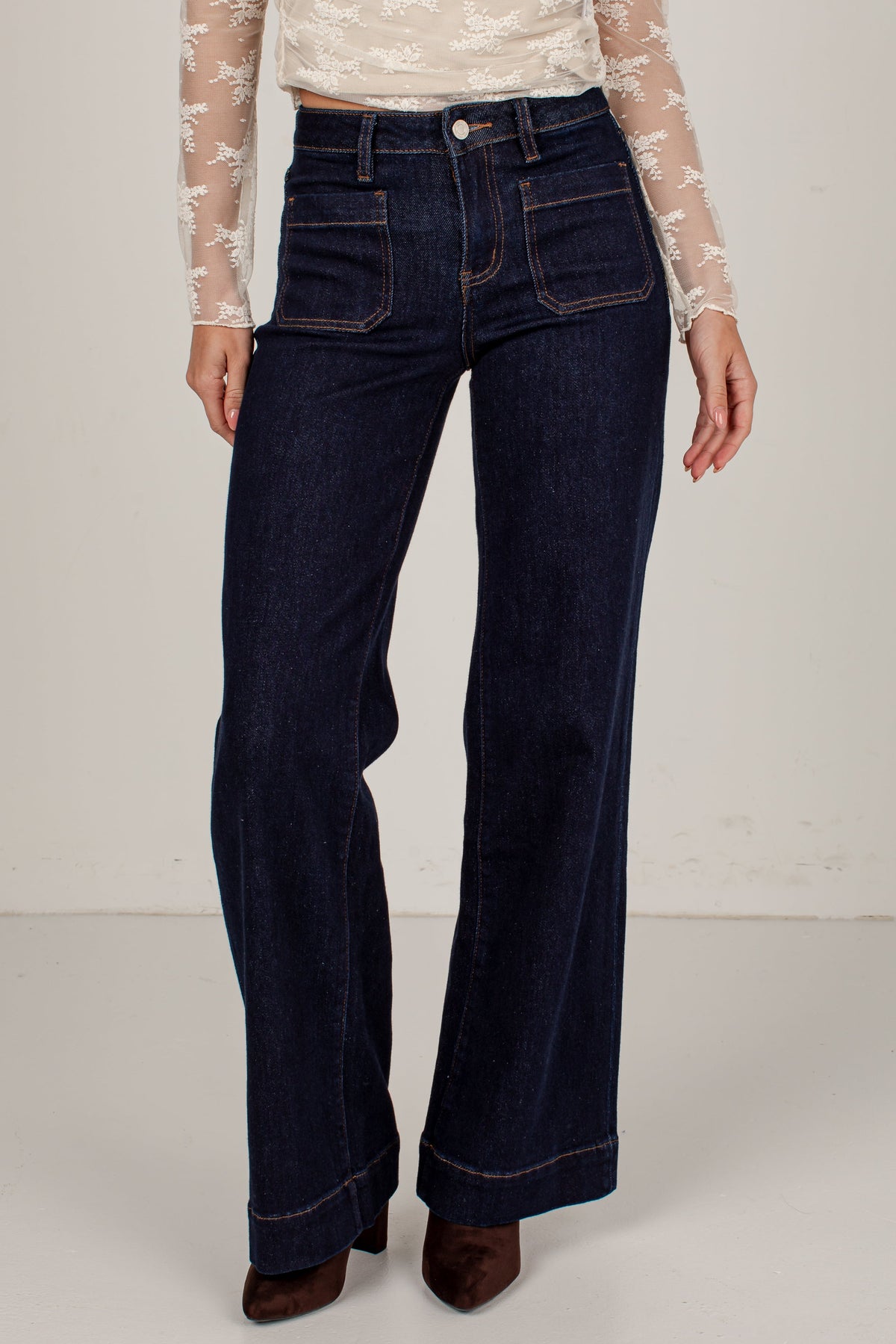 A model wearing dark blue wide-leg denim jeans with a beige lace long-sleeve top and brown suede boots, shown from the waist down against a light off-white studio background.