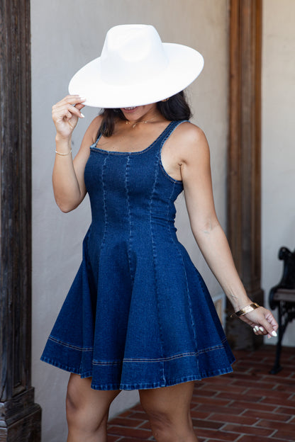 A woman wearing a blue denim dress and a large white wide-brim hat, standing in a doorway with a brick floor.