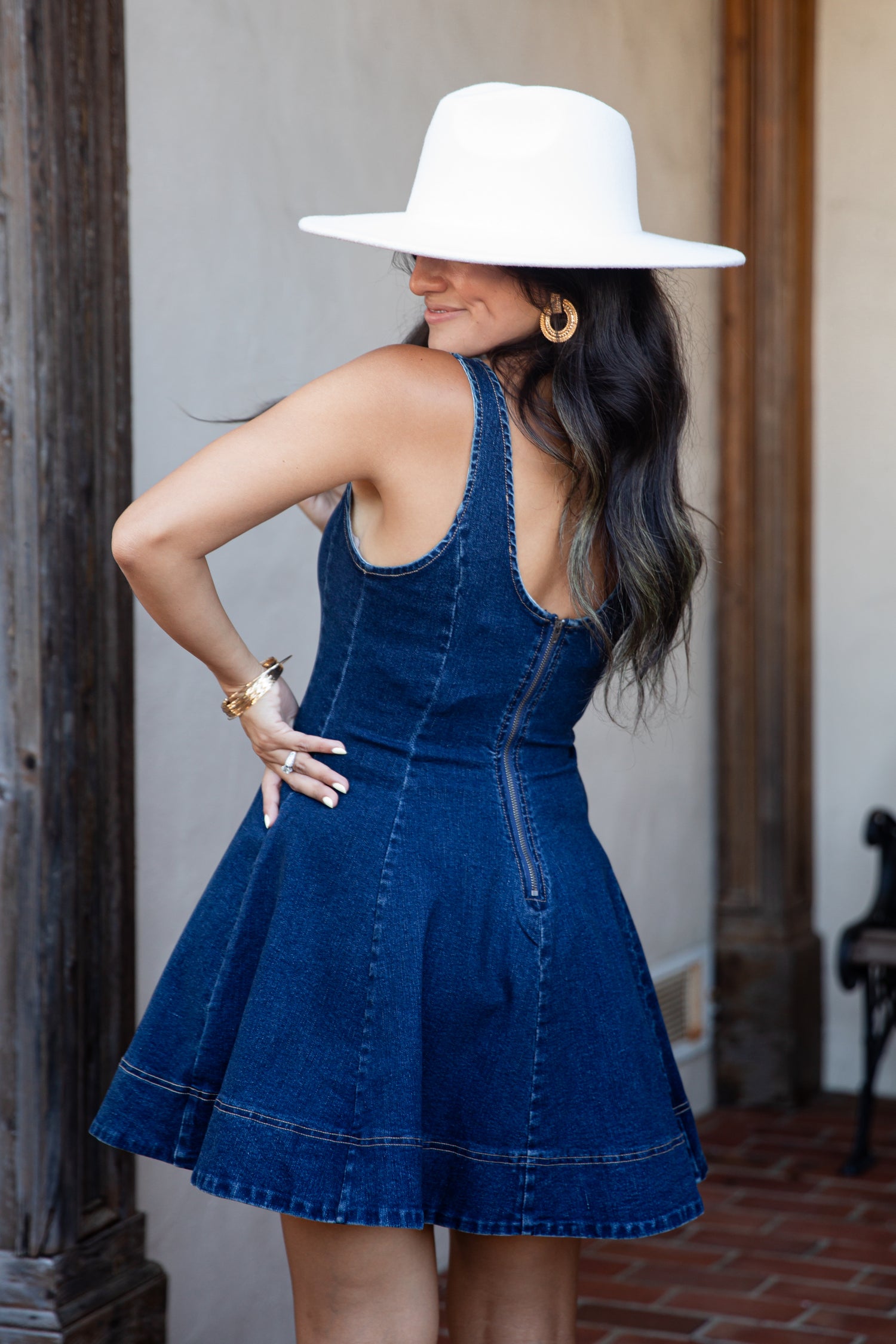A woman wearing a dark blue denim pinafore dress with a flared skirt, styled with a wide-brimmed white hat, gold hoop earrings, and a gold bracelet, posing in front of a rustic wooden door and stucco wall.