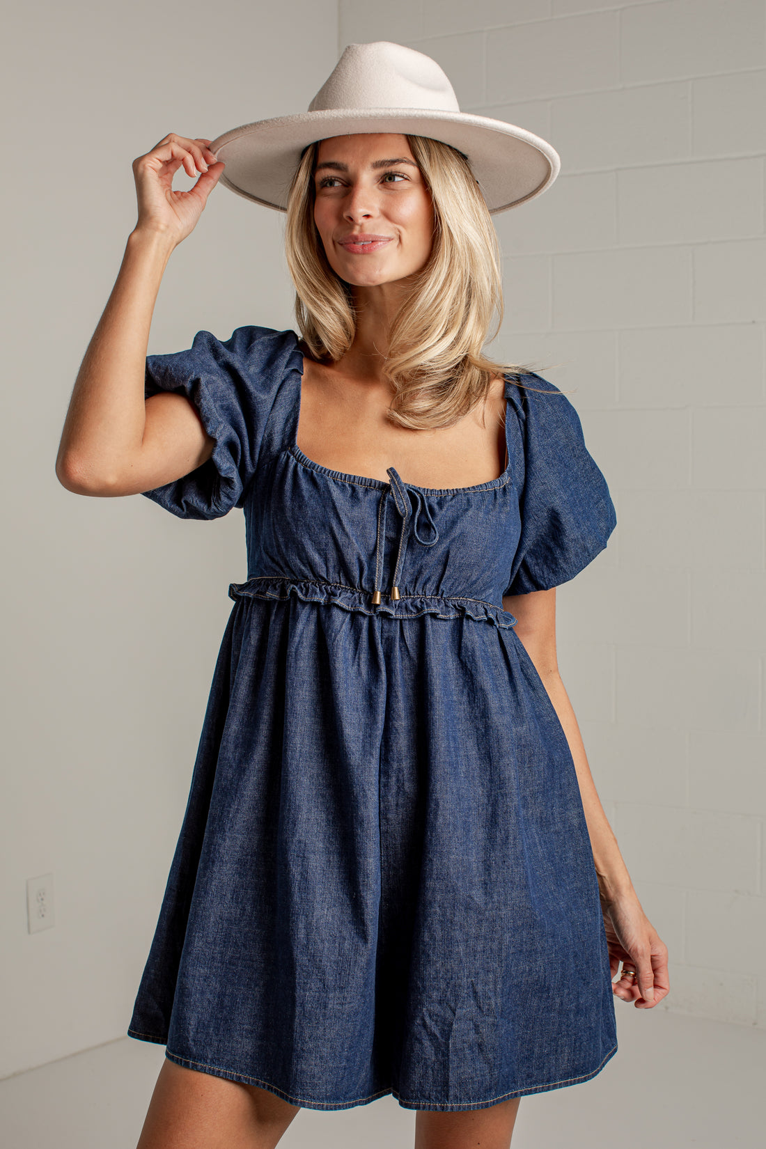 A woman wearing a blue denim dress with puff sleeves, a square neckline, and a drawstring waist, paired with a wide-brimmed white hat, posing against a light gray wall.