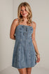 A woman modeling a light blue denim mini dress with spaghetti straps and a lace-up front, standing against a white brick wall.