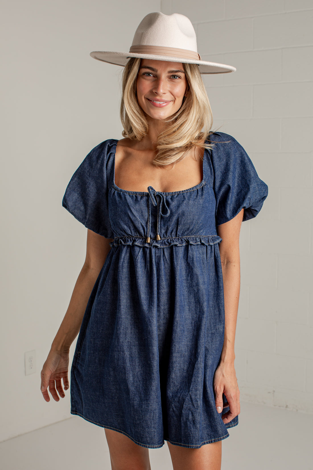 A woman wearing a blue denim mini dress with puff sleeves, a square neckline, and a drawstring tie, paired with a wide-brimmed cream hat, standing against a light gray brick wall.