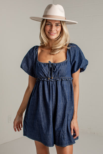 A woman wearing a blue denim mini dress with puff sleeves, a square neckline, and a drawstring tie, paired with a wide-brimmed cream hat, standing against a light gray brick wall.
