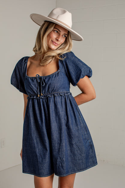 A woman wearing a blue denim romper dress with puff sleeves, a square neckline with a tie detail, and a gathered waist, paired with a wide-brimmed cream hat, standing against a light gray brick wall.