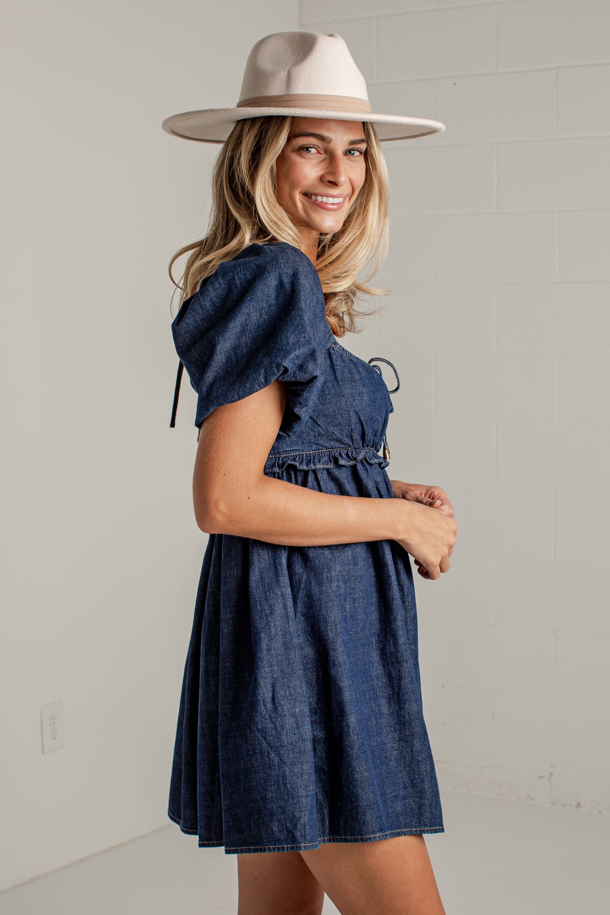 A woman wearing a dark blue denim puff-sleeve dress and a beige wide-brim hat, posing against a light gray brick wall.