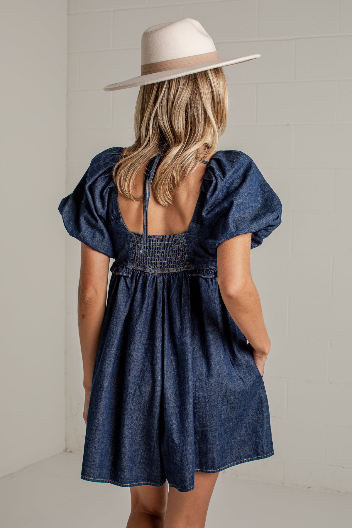 A woman viewed from behind wearing a dark blue denim-like puff-sleeve dress with a smocked back and a cream wide-brim hat, standing against a light neutral wall.