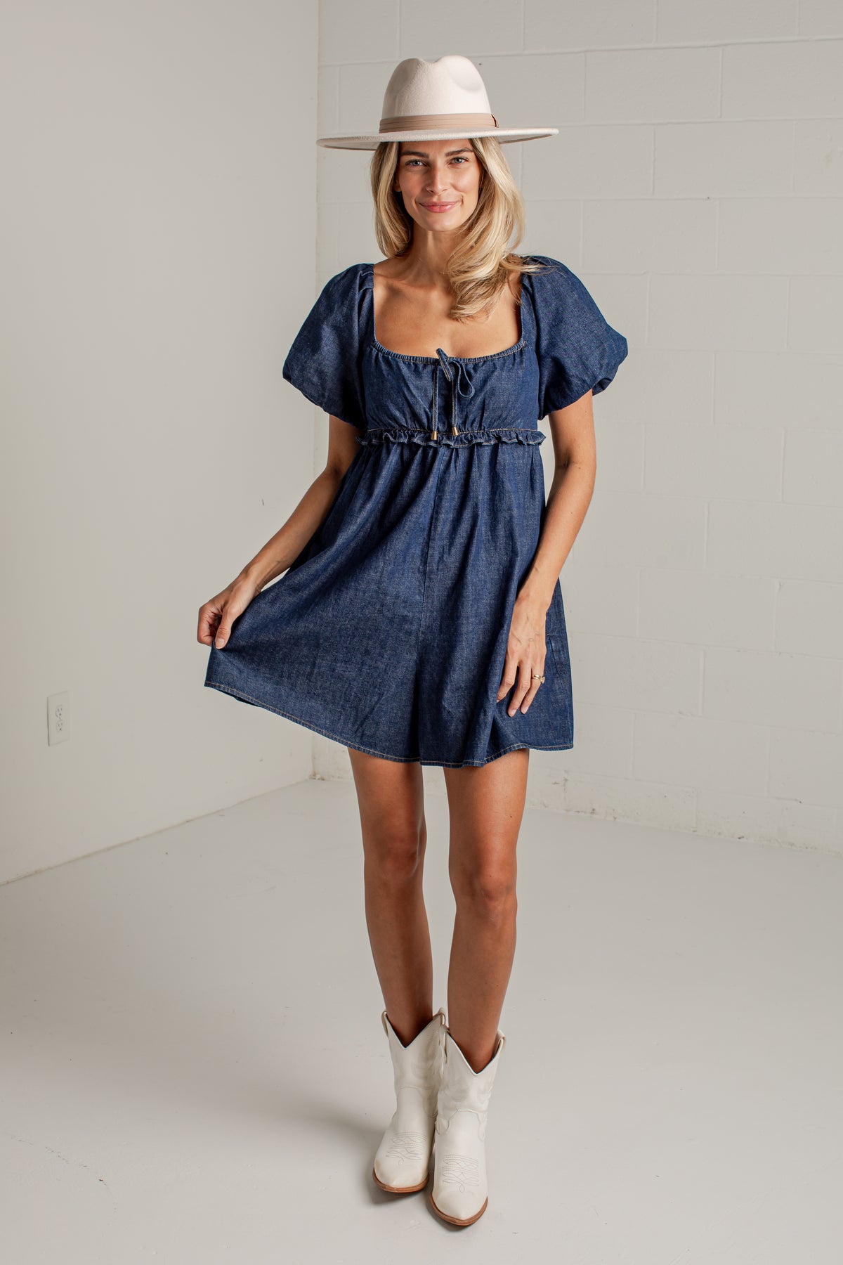 A woman modeling a short blue denim dress with puff sleeves, a square neckline, and a gathered waist, styled with a beige wide-brim hat and white cowboy boots in a bright studio setting.