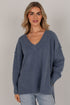 A woman wearing an oversized, heathered slate blue V-neck knit sweater with light-wash jeans, standing against a neutral light gray studio background.