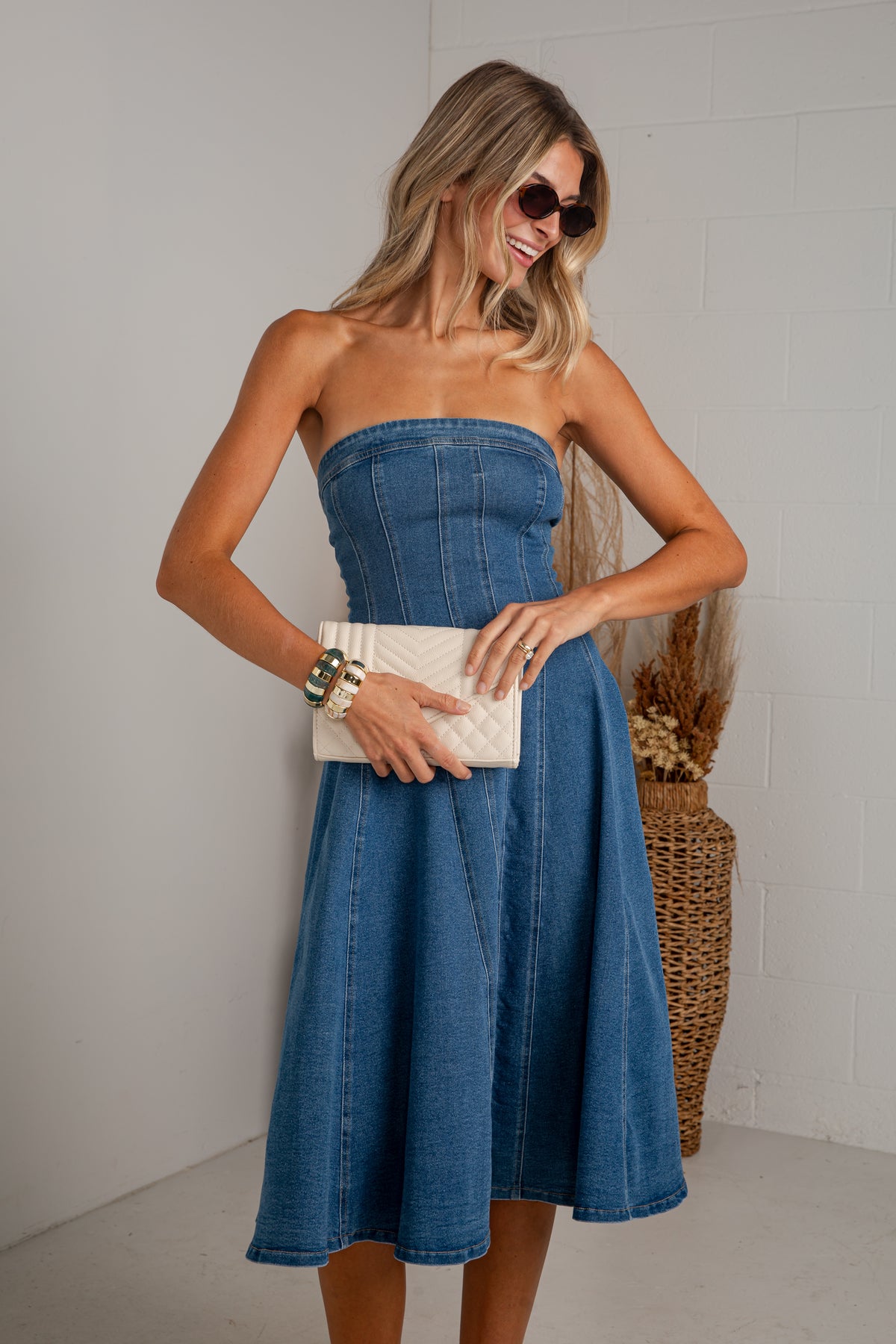 A woman modeling a strapless blue denim midi dress with a corset-style bodice and flared skirt, holding a cream quilted clutch against a light gray wall.