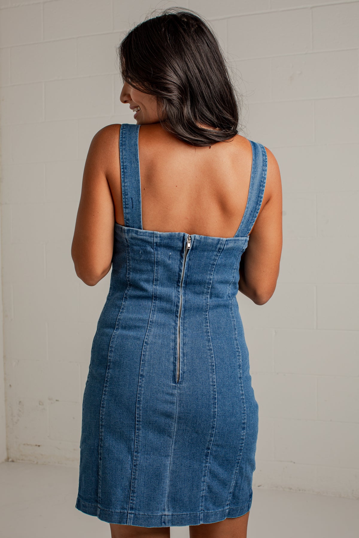 A woman wearing a blue denim mini dress, shown from the back against a white brick wall.