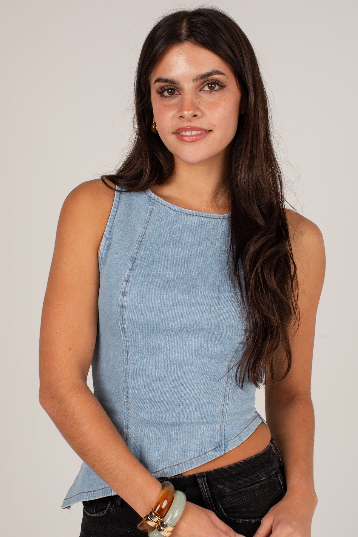 A woman wearing a light blue sleeveless cropped top with a curved hem, paired with black jeans, against a plain light gray studio background.