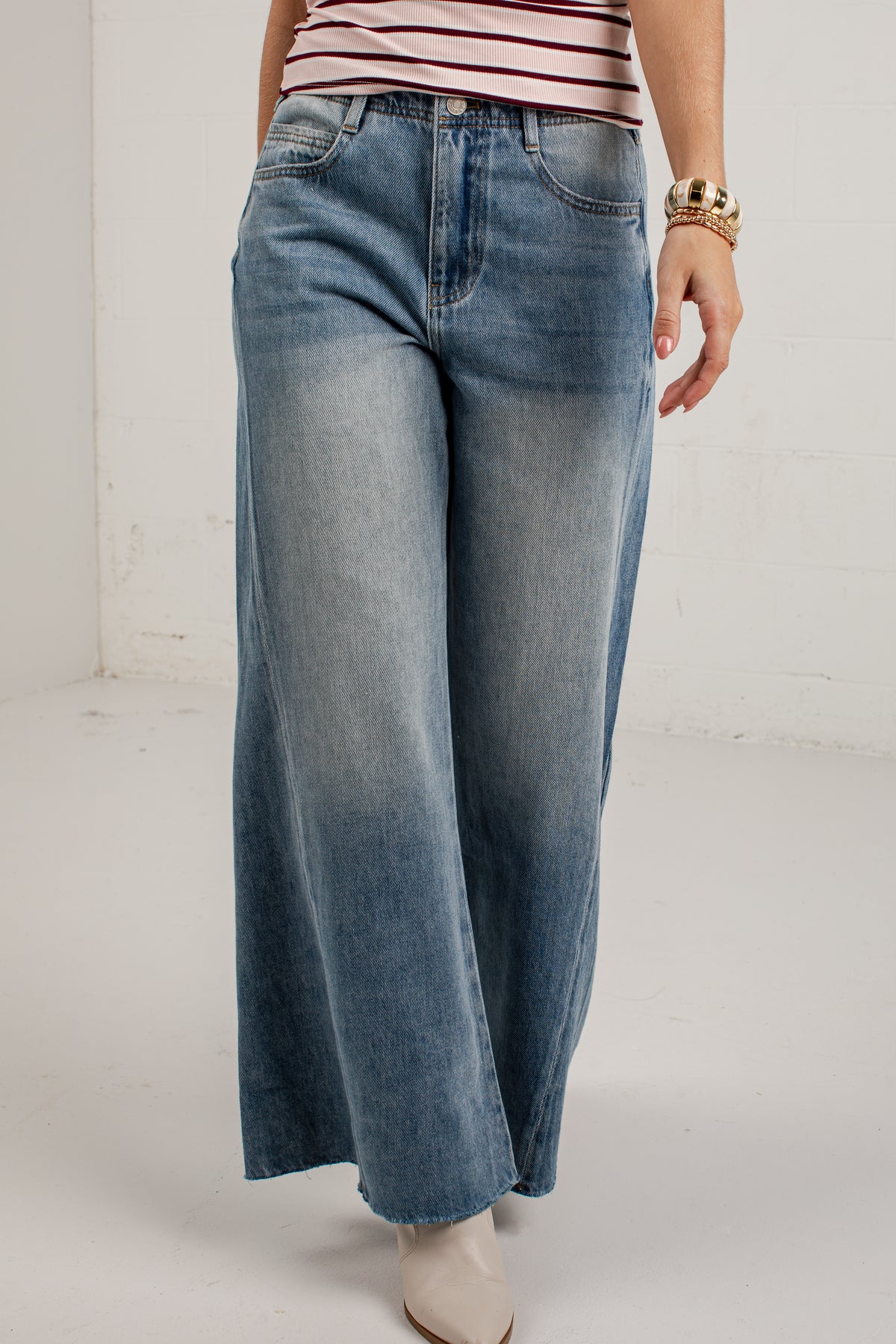 A person wearing wide-leg blue denim jeans with a faded wash, paired with a pink and white striped top and beige ankle boots, standing against a plain light gray wall.