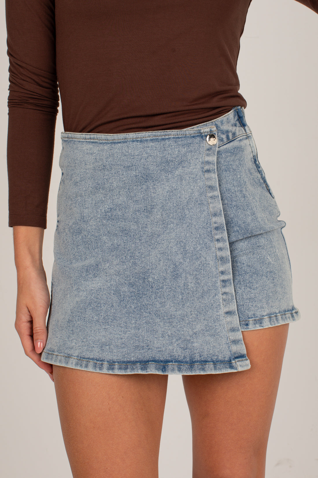 A person wearing a light-wash denim wrap-style mini skirt with a brown long-sleeve top, shown from the chest to mid-thigh against a plain light beige studio background.