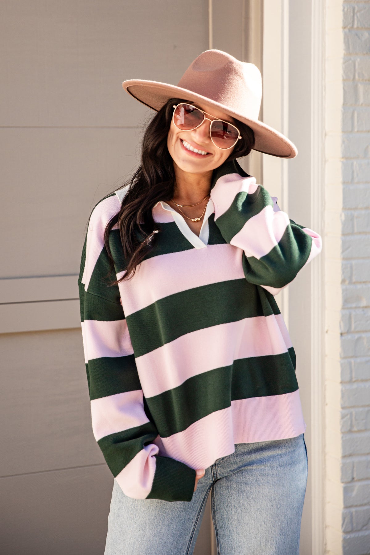 A woman wearing a pink and dark green horizontally striped long-sleeve knit top, light-wash blue jeans, a dusty pink wide-brim hat, and sunglasses, standing outdoors against a beige paneled wall.