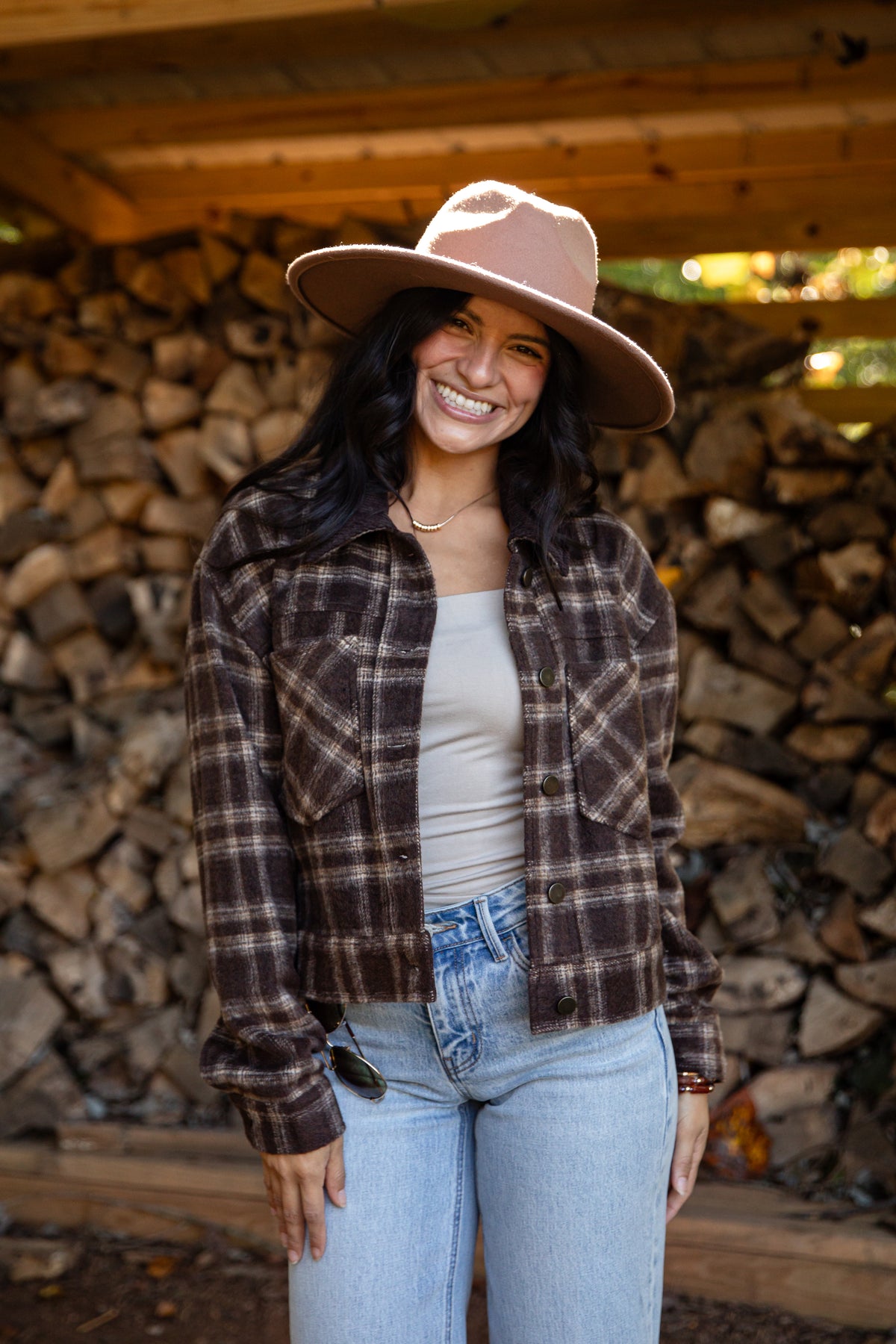 A woman smiling in front of a stacked woodpile, wearing a brown and black plaid flannel shirt jacket, a beige tank top, light blue jeans, and a dusty pink wide-brim hat.