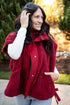 A woman wearing a burgundy sherpa fleece vest over a matching red button-front jacket, styled with a white long-sleeve shirt and light blue jeans, standing outdoors in a sunny park setting.