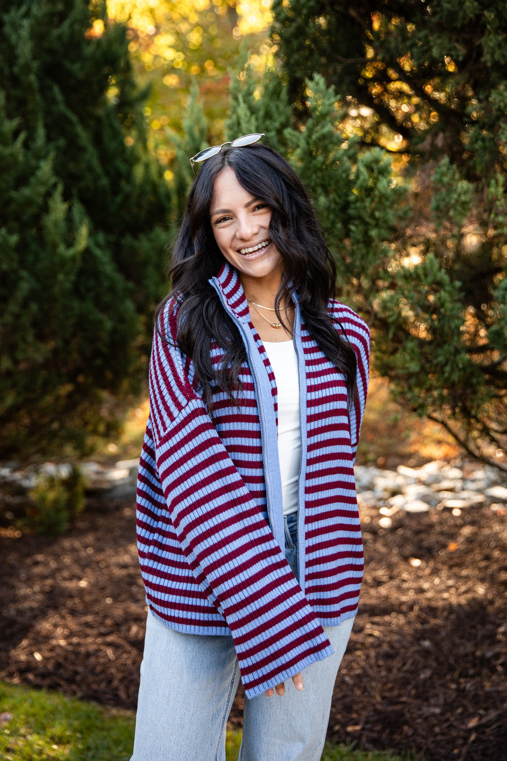 A woman smiling outdoors wearing a red, white, and blue striped knit poncho-style cardigan over a white top and light blue jeans, with trees and mulch in the background.