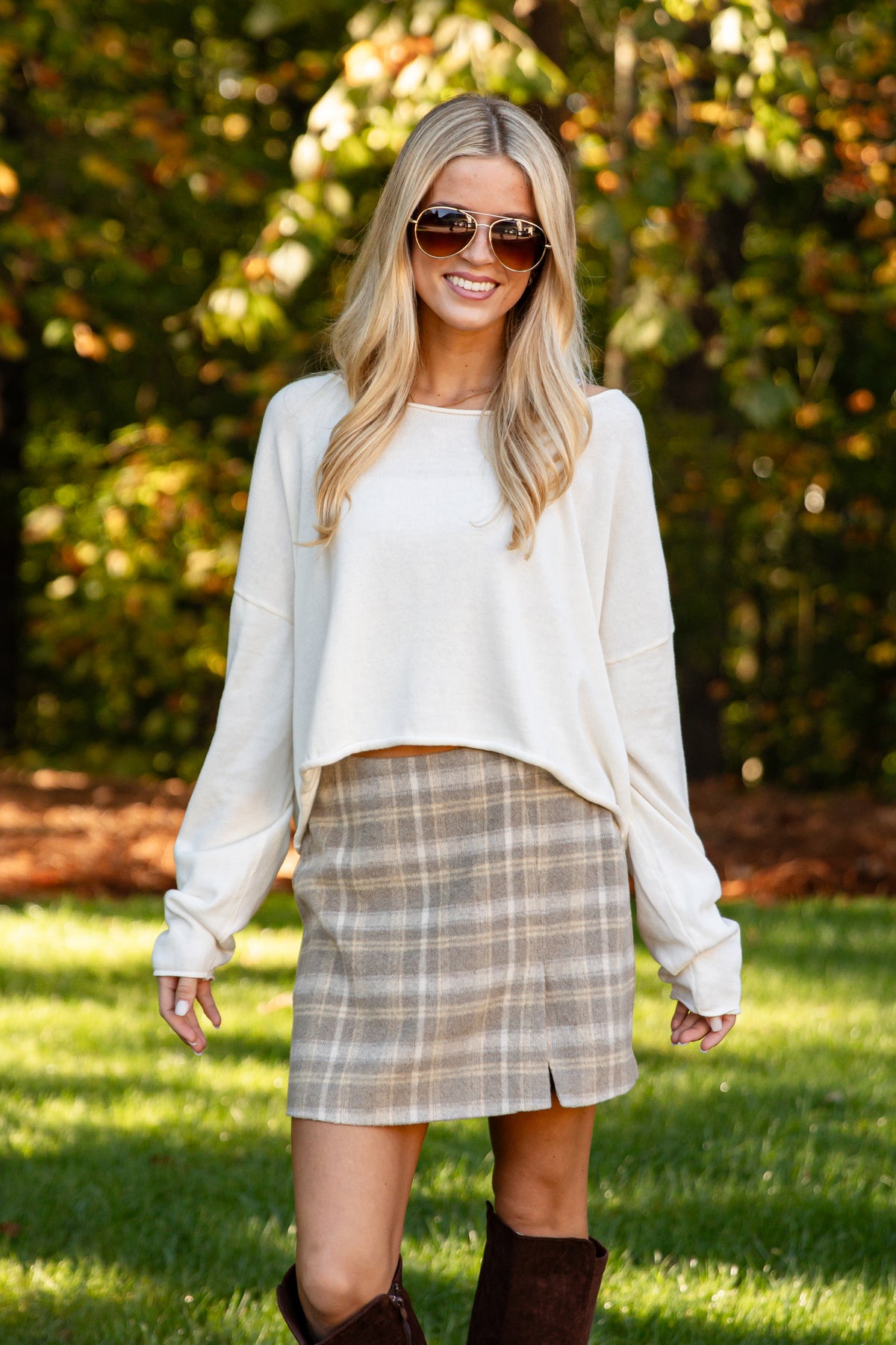 A woman wearing a cream long-sleeve cropped sweater and a beige and brown plaid mini skirt, standing outdoors on a grassy lawn with trees in the background.