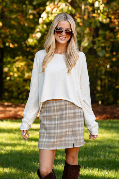 A woman wearing a cream long-sleeve cropped sweater and a beige and brown plaid mini skirt, standing outdoors on a grassy lawn with trees in the background.