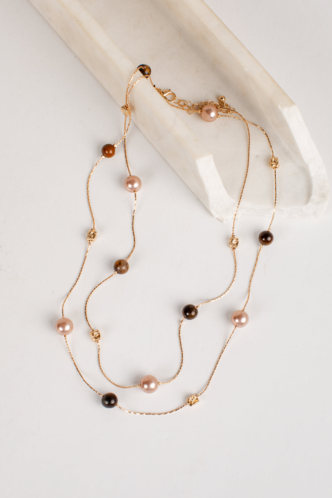 A double-strand gold chain necklace featuring alternating pink freshwater pearls, brown pearls, and small gold spacer beads, displayed on a light cream marble prop against a white background.