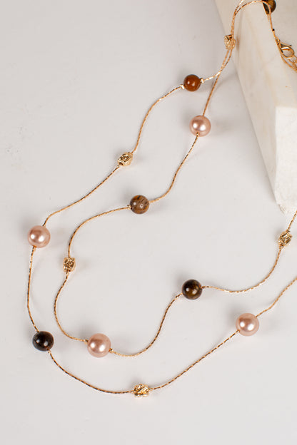 A gold-toned multi-strand necklace featuring faux pearls in light pink, dark brown, and black, accented with small gold beads, displayed on a light gray surface next to a white marble prop.