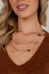 A close-up of a woman wearing a double-layered necklace with gold chains, small gold beads, and alternating pearl and dark brown round beads, paired with a brown knit sweater.