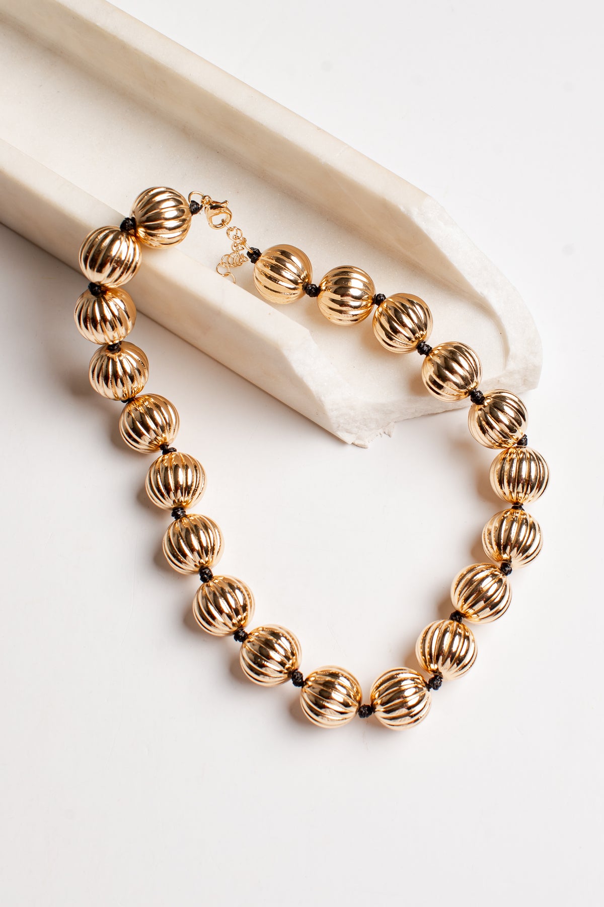 A gold-toned beaded necklace with ribbed, textured spherical beads and small dark spacer beads, displayed on a cream fabric prop against a white background.